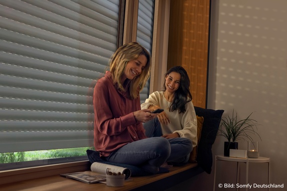 Zwei junge Frauen sitzen auf der Fensterbank und schließen die Rollläden per Handy