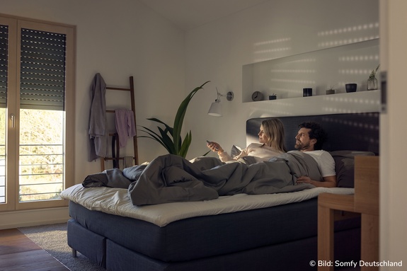 Paar öffnet Rollläden aus dem Bett per Fernbedienung