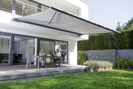 Helle Markise über Terrasse eines modernen Hauses, darunter speisen zwei Personen.