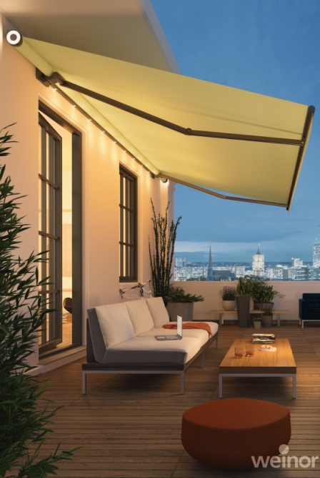 Moderne Terrasse abends, warm beleuchtet, mit ausgefahrener heller Markise, Sofa und Stadtskyline.