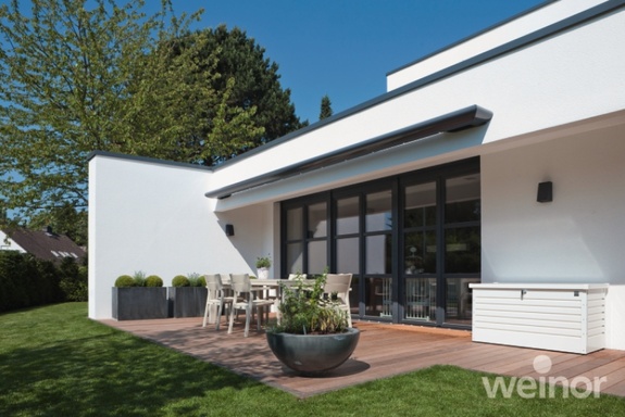 Modernes weißes Haus mit Holzterrasse, Gartenmöbeln, grünem Rasen und blauem Himmel.
